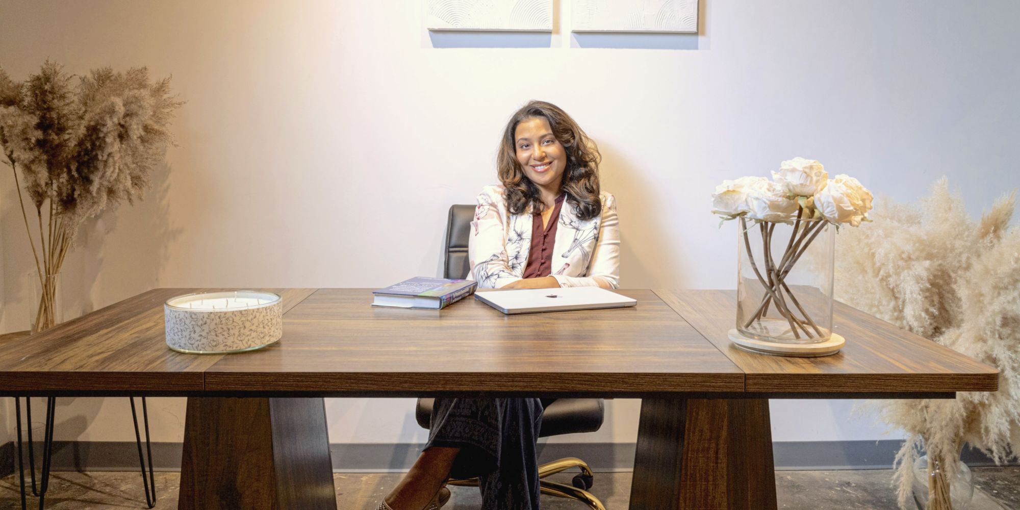 Hoda Ammar smiling at a wooden desk in a calm, softly lit room with white flowers and pampas grass